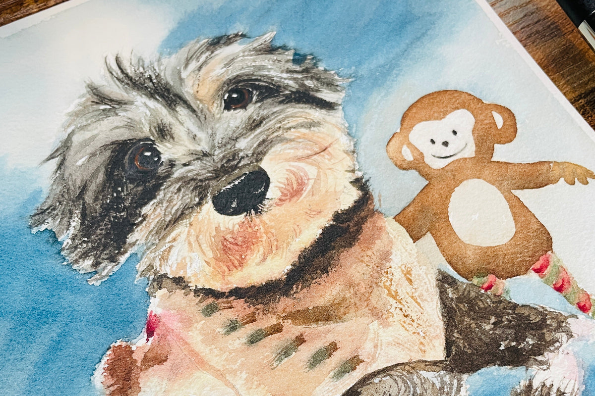 A watercolor painting of a fluffy gray and black dog wearing a pink sweater, with a small brown monkey figure beside it, shown on a white sheet of paper over a wooden surface.