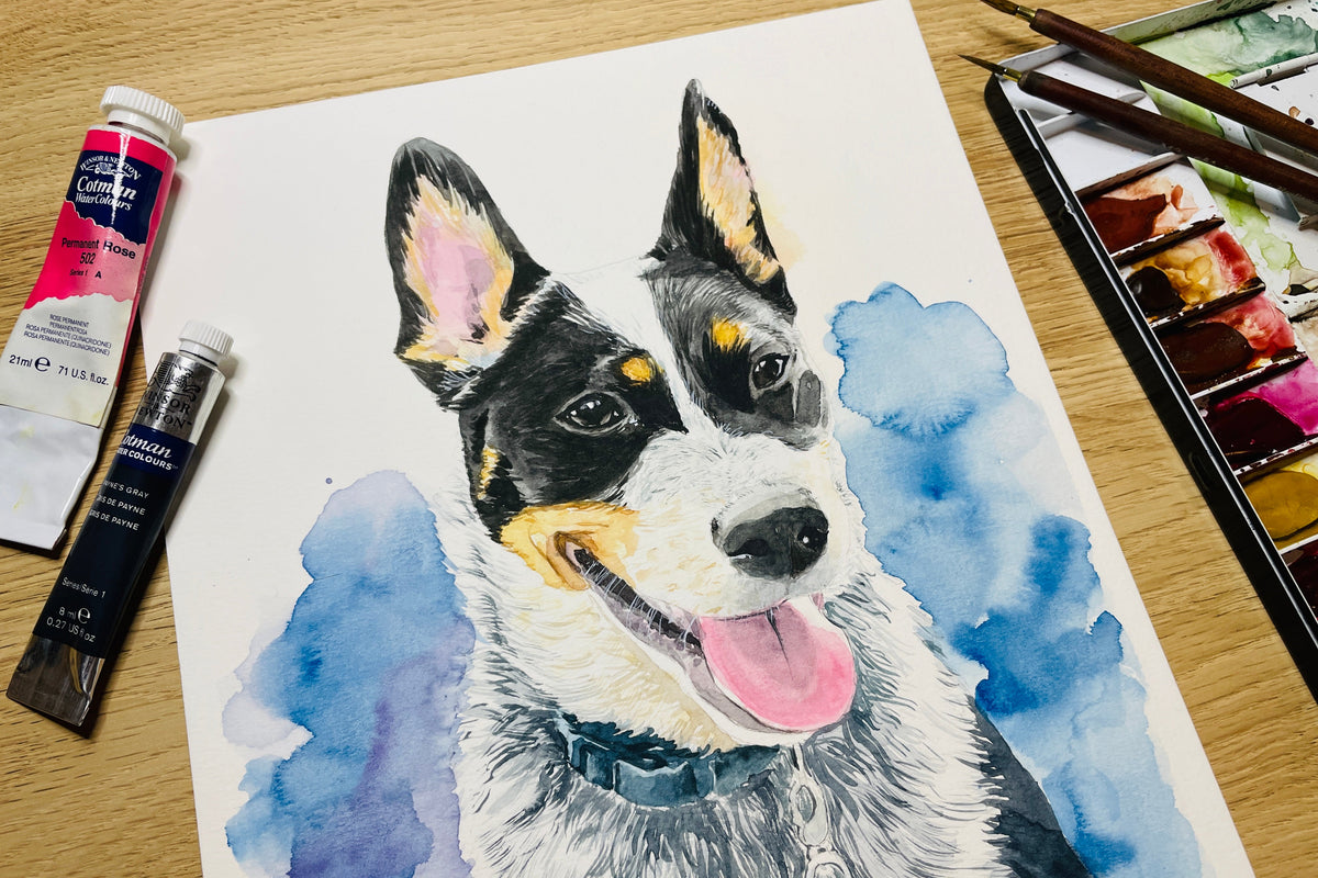A watercolor painting of a black, white, and tan dog with its tongue out, set against blue watercolor washes on a sheet of paper. The paper is displayed on a wooden table alongside an artist's paint tube, paintbrushes, and a watercolor palette.