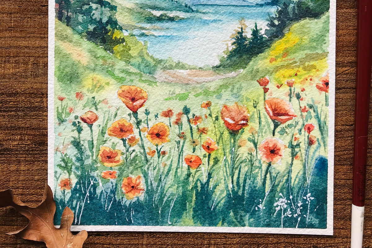 A watercolor landscape painting on textured paper, depicting a scenic valley with mountains, pine trees, a lake, and a field of red poppies, displayed on a wooden surface with art tools.